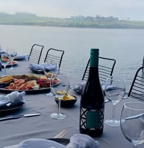 Outdoor dining table set with plates, wine glasses, a bottle of Waterton Hall wine, and a platter of food, overlooking a body of water and distant landscape.
